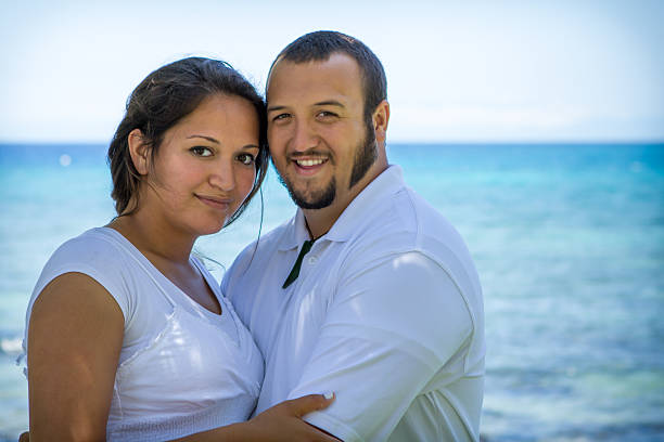 Couple hugging and smiling towards the camera as they stand in front of the ocean.