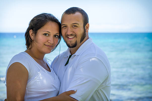 Couple hugging and smiling towards the camera as they stand in front of the ocean.