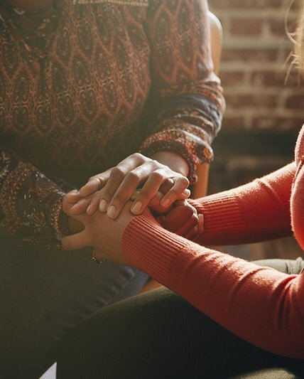 close up of two seated women's clasped hands with sun streaming through a window.