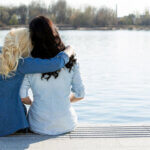 Two women sitting on a dock, one with her head leaning on the other with her arm around her friend, staring out at a lake.