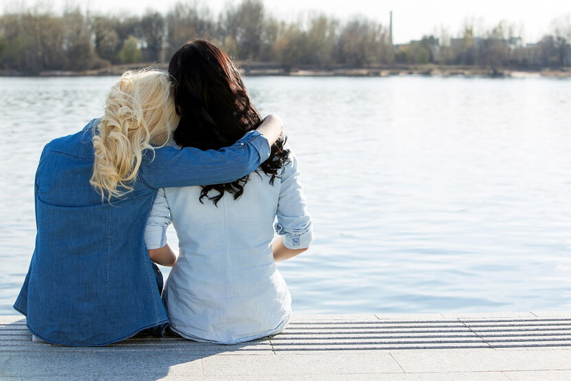 Two women sitting on a dock, one with her head leaning on the other with her arm around her friend, staring out at a lake.