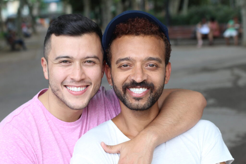 A LGBTQ+ couple, one with his arm around his partner's shoulder, holding his chest, who stands slightly in front of him, smiles widely as they face the camera.