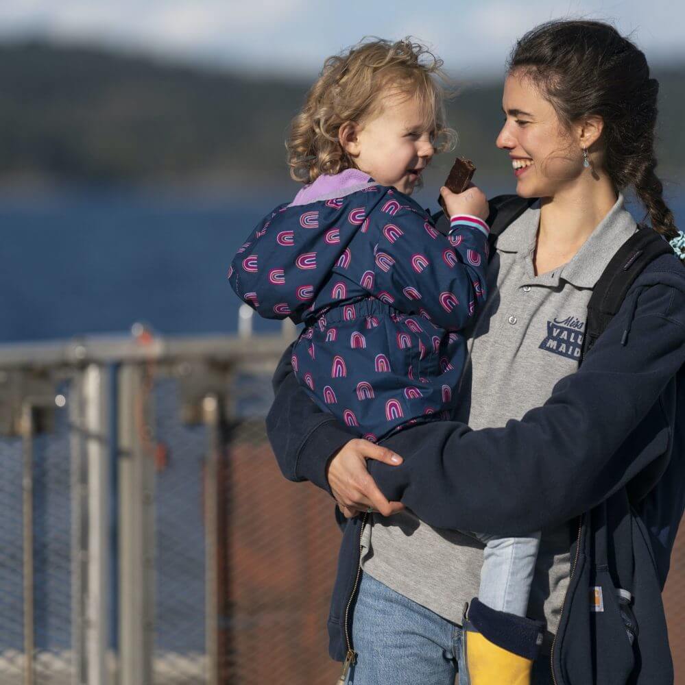 Still shot of the characters Alex carrying Maddy, who play mother and daughter in the movie MAID, as they smile, standing in front of a metal fence and body of water.